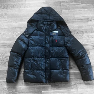 Svart pufferjacka från Polo Ralph Lauren - Svart pufferjacka från Polo Ralph Lauren med huva, dragkedja och tryckknappar. Jackan har det klassiska röda Polo-logot broderat på bröstet och är tillverkad i glansig syntet. Perfekt för kalla dagar med sin quiltade design och snygga passform.