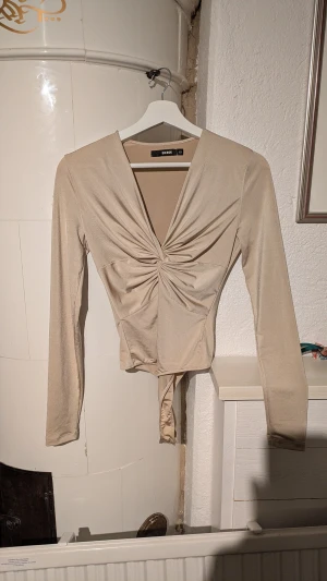 Beige omlottopp från Bik Bok - Säljer en beige omlottopp från Bik Bok i storlek XS. Toppen har lång ärm, snygg v-ringning och cool twistad detalj framtill som ger en unik look. Materialet är mjukt och har lite glans, perfekt för att sticka ut. Passar dig som gillar stilrena och trendiga plagg.