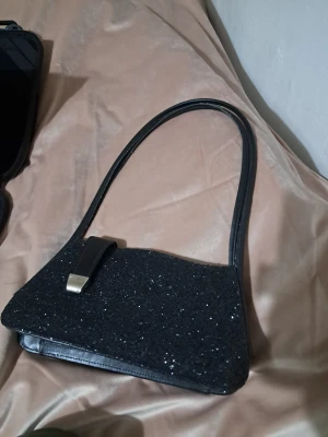 Svart glittrig handväska med skinnrem - Snygg svart handväska med glittrig yta och stilren form. Väskan har en lång, smal axelrem i svart skinn och ett dekorativt spänne i silverfärg. Perfekt för dig som vill sticka ut med en elegant accessoar. Lite skador se bilder