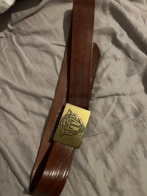 Brunt skärp från Tiger of Sweden - Snyggt brunt bälte i skinn från Tiger of Sweden med en bred, rektangulär mässingsfärgad spänne och präglad logga. Bältet har diskreta längsgående ränder och en klassisk look som sticker ut med sitt coola spänne. Använder inte längre därför säljs det