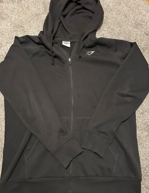 Svart hoodie från Gymshark med dragkedja - Svart hoodie från Gymshark i storlek M med hel dragkedja och huva med snörning. Tröjan har ribbade muddar, fickor framtill och en diskret Gymshark-logga på bröstet. Materialet är en mix av bomull och polyester, vilket gör den mjuk och skön.