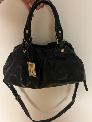 Svart handväska från Marc Jacobs - Snygg svart handväska från Marc Jacobs i mjukt skinn med guldfärgade detaljer och dragkedja. Väskan har både handtag och axelrem, samt ett coolt innerfoder med grafiskt mönster och logga. Perfekt storlek för att få plats med allt du behöver. Lite defekter på handtagen, se bild 3, men syns knappt.