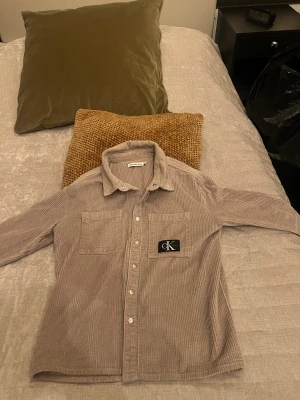 Beige manchester skjorta Calvin Klein - Snygg beige skjorta i mjuk manchester från Calvin Klein Jeans. Skjortan har två bröstfickor, klassisk krage och vita knappar framtill. På ena fickan sitter en svart CK-logga som ger extra stilpoäng. Perfekt för dig som gillar en chill och trendig look.