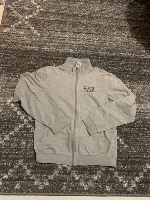 Grå ziptröja EA7 Emporio Armani - Snygg grå ziptröja från EA7 Emporio Armani med hög krage och diskret logga på bröstet. Tröjan har hel dragkedja, två fickor fram och ribbade muddar vid ärmslut och nederkant. Perfekt för chill eller träning, stilren och enkel att matcha.