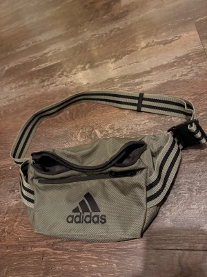Grå axelväska från Adidas - Snygg grå axelväska från Adidas med svart logga framtill. Väskan har ett stort fack med dragkedja och en mindre ficka på framsidan. Justerbart randigt band i svart och grått gör den enkel att bära över axeln. Perfekt för att ha med sig det viktigaste. Oanvänd