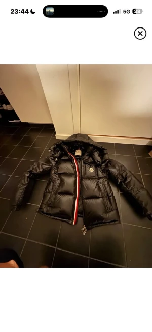 Svart pufferjacka från Moncler - Svart pufferjacka från Moncler med glansig finish och ikonisk logga på bröstet. Jackan har huva, röda och vita detaljer vid dragkedjan samt två stora fickor framtill. Perfekt för kalla dagar och har en riktigt snygg streetstil.