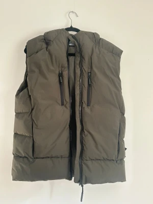 Olivgrön dunväst från Stone Island - Snygg olivgrön dunväst från Stone Island med ikonisk patch på sidan. Västen har två vertikala dragkedjefickor framtill och en rejäl dragkedja i mitten. Ingen CLG kod finns.