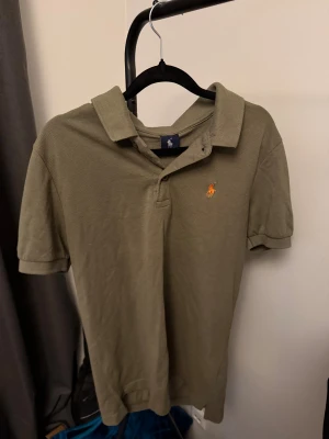Olivgrön pikétröja från Polo Ralph Lauren - Olivgrön pikétröja från Polo Ralph Lauren med klassisk krage och korta ärmar. Tröjan har den ikoniska broderade loggan i orange på bröstet och är gjord i mjuk bomull. Perfekt för dig som gillar stilrena och tidlösa plagg.