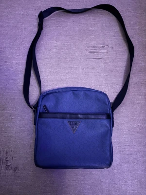 Blå axelväska från Guess ÄKTA - Snygg blå axelväska från Guess med justerbar svart rem och diskret mönster av små G:n över hela väskan. Väskan har dragkedja upptill, ett ytterfack med dragkedja och flera innerfack. Perfekt storlek för det viktigaste och stilren design med Guess-logga framtill.