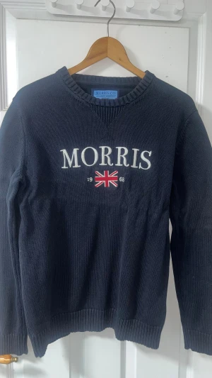 Mörkblå stickad tröja Morris  - Snygg mörkblå stickad tröja från Morris Co i storlek medium. Tröjan har vit broderad logga och brittisk flagga framtill, samt ribbade muddar vid hals, ärmslut och nederkant. Perfekt för dig som gillar klassisk stil med sportig touch.