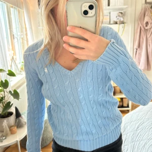 Ljusblå kabelstickad tröja Ralph Lauren - Ljusblå kabelstickad tröja från Ralph Lauren Sport i 100% bomull. V-ringad modell med vit broderad logga på bröstet. 