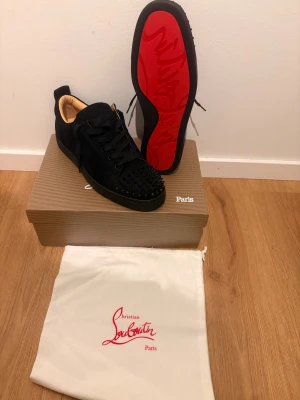 Christian Louboutin svarta sneakers med nitar - Svarta sneakers från Christian Louboutin i mocka med ikonisk röd sula och svarta nitar på tån. Skorna har snörning och rund tå, samt lyxig beige insida. Perfekt för dig som vill sticka ut med en edgy och exklusiv look.