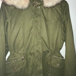 Olivgrön parkas med riktigt päls  - Snygg olivgrön parkas med huva och fluffig fuskpäls runt kragen. Jackan har dragsko i midjan, stora fickor med dragkedja och tryckknappar. Perfekt för kyliga dagar och har en klassisk, avslappnad look.