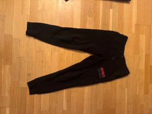 Svarta mjukisbyxor från Hoodrich M - Svarta mjukisbyxor från Hoodrich med tryckt logga i rött och vitt på ena benet. Byxorna har snörning i midjan, ribbade muddar vid bensluten och en bakficka. Perfekta för chill eller streetwear-stil.