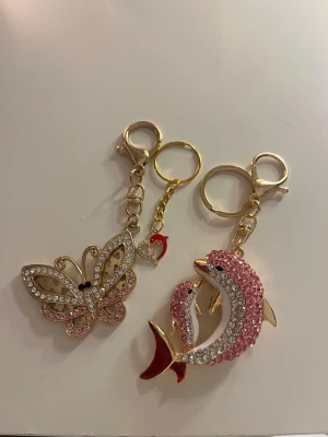 Nyckelringar med fjäril och delfiner -  glittriga nyckelringar i guldmetall, en med en fjäril dekorerad med rosa och vita strasstenar, och en med två delfiner täckta av rosa och vita stenar samt röd detalj på stjärtfenan. Perfekt som accessoar till väskan eller nycklarna.