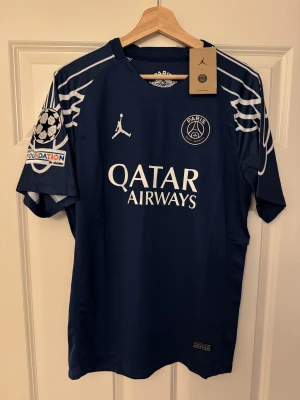 PSG Ousmane Dembélé matchtröja blå - Säljer en officiell PSG-tröja med Ousmane Dembélé #10. Marinblå med vita detaljer, kortärmad och med snygga grafiska linjer på axlarna. Jordan-logga och Qatar Airways-tryck. Lätt polyester.  Följ oss gärna på Instagram: @kickstoreuf.