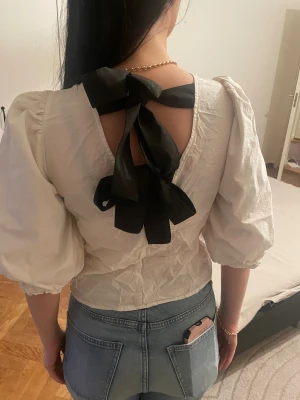 Vit blus med puffärm och svart rosett - Supersöt vit blus med puffiga trekvartsärmar och en stor svart rosett i ryggen. Blusen har öppen rygg och är gjord i ett lätt, krispigt bomullstyg. Perfekt för dig som gillar statement-detaljer och vill sticka ut lite extra. Snygg att styla med jeans eller kjol. Passar även S😊Obs stryker innan köp💕