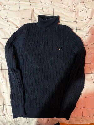 Mörkblå polotröja från GANT - Snygg mörkblå polotröja från GANT med klassisk kabelstickad design och broderad logga på bröstet. Tröjan har lång ärm och hög polokrage som ger en chill och stilren vibe. Perfekt för dig som gillar preppy stil och vill ha något varmt och mjukt.