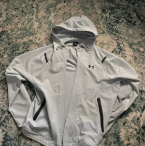  vindjacka från Under Armour helt Ny - Snygg vit vindjacka från Under Armour med huva och svarta detaljer vid dragkedjorna. Jackan har två sidofickor med dragkedja och en sportig look, perfekt för dig som gillar streetstyle. Justerbar snörning vid huvan för extra komfort.
