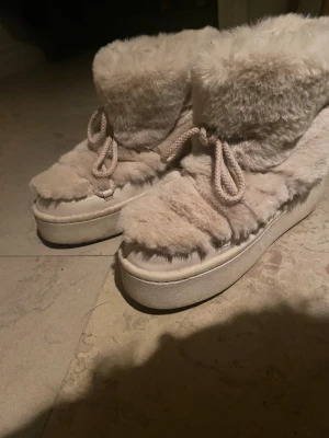 Beige fluffiga skor med snörning - Supermysiga vinterskor i beige med fluffigt pälsmaterial över hela skon och tjock platt sula. De har snörning framtill och en rund tå. Kan matchas supergulligt med fluffiga vantar och en mössa.