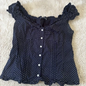 brandy melville topp - Supersöt mörkblå blus från Maison Scotch med vita prickar, volangkant vid axlar och nederkant samt knappar framtill. Blusen har en feminin och luftig passform, perfekt för dig som gillar romantiska detaljer och klassiskt mönster.
