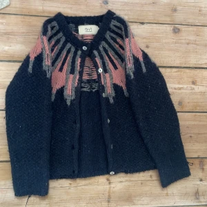 Mönstrad kofta från Emma och Malena - Mysig stickad kofta i mörkblått från Emma och Malena med unikt mönster i rosa och grått upptill. Koftan har knappar framtill, lång ärm och en riktigt skön, lite oversized passform. Perfekt för kalla dagar när du vill ha något både snyggt och bekvämt.