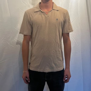 Pikétröja från Morris - Beige pike från Morris med klassisk krage och v-ringning utan knappar. Piken har korta ärmar och en liten  broderad logga på bröstet. Tillverkad i mjuk bomull som känns skön mot huden. Perfekt för en clean och stilren look. Modellen är 180cm