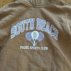 Brun hoodie South Beach Padel - Brun hoodie från South Beach med tryck 'South Beach Padel Sports Club' och motiv av padelracketar i vitt och lila. Tröjan har huva, ribbade muddar och en avslappnad passform. Perfekt för dig som gillar sportig stil.