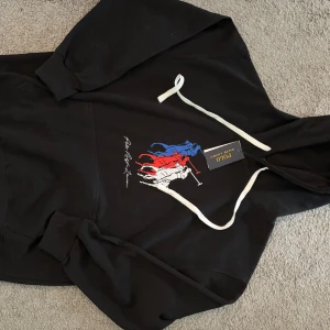 Svart hoodie Polo Ralph Lauren M - Svart hoodie från Polo Ralph Lauren med vit dragsko och stort tryck framtill i blått, rött och vitt som föreställer polospelare. Klassisk känguruficka och ribbade muddar. Perfekt för dig som gillar streetwear och snygga loggor.