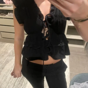 Svart volangblus med snörning - Supersöt svart blus med volanger och snörning framtill. Toppen har korta ärmar, djup v-ringning och croppad passform. Perfekt för dig som gillar trendiga detaljer och en feminin look.