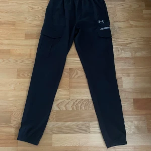 Svarta cargobyxor från Under Armour - Svarta cargobyxor från Under Armour med elastisk midja och stora fickor på sidorna. Byxorna har en sportig look, smal passform och är tillverkade i ett lätt syntetmaterial. Perfekta för dig som gillar streetwear och funktionella detaljer.