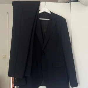 Svart kostym från Riley Slim Fit - Säljer en stilren svart kostym från Riley i slim fit-modell. Kavajen har klassisk passform med två knappar, fickor med lock och slät design. Byxorna är matchande svarta och raka. Perfekt för dig som vill ha en enkel och snygg look. Nypris 4500 säljer ätt sätt både byxor och kostymen storlek på båda 152=L säljer för 1000kr