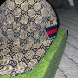 Gucci keps med GG-mönster, beige/blå - Säljer en snygg Gucci keps i beige och blått med klassiskt GG-monogram över hela kepsen. Kepsen har justerbart spänne bak i blått och en röd/blå rand på sidan. Tillverkad i bomullscanvas och detaljer i skinn. Perfekt för dig som gillar lyxiga accessoarer.