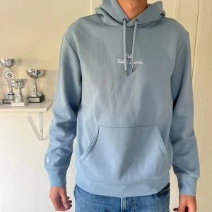 Ljusblå hoodie från Polo Ralph Lauren - Säljer en helt ny ljusblå hoodie från Polo Ralph Lauren med broderad logga på ärmen och vit text på bröstet. Tröjan har huva med snörning och är tillverkad i mjuk bomull. Storlek M, 1100kr