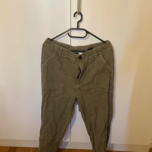 Olivgröna cargobyxor från H&M - Snygga olivgröna cargobyxor från H&M med stora fickor framtill och markerade sömmar. Byxorna har en avslappnad passform och är tillverkade i bomull, perfekta för en chill och trendig look.