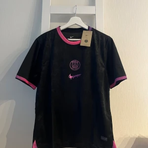 PSG x Nike svart fotbollströja M - Svart PSG fotbollströja från Nike med rosa och lila detaljer på krage och ärmslut. Paris Saint-Germain-logga och unik rosa Nike swoosh framtill. Tillverkad i lätt Dri-FIT-material som andas. Perfekt för dig som vill sticka ut på planen.