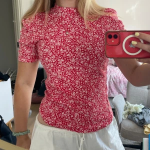 Röd blommig topp från Kappahl - Superfin röd topp från Kappahl med vitt blommönster och ribbad struktur. Modellen är figurnära med korta ärmar och hög hals. Perfekt för dig som gillar färg och mönster! Materialet är mjukt och stretchigt, vilket gör den riktigt bekväm att bära.