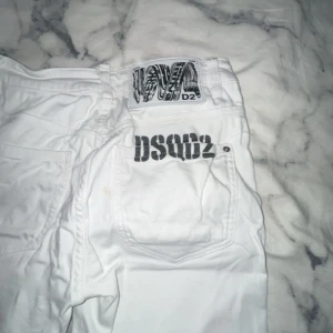 Vita jeans från Dsquared2 - Säljer ett par vita jeans från Dsquared2 med coola slitningar på benen och tydlig logga på bakfickan. Klassisk femficksmodell med röd Dsquared2-tag vid fickan. Snygg passform och stilren look, perfekta för dig som vill sticka ut.