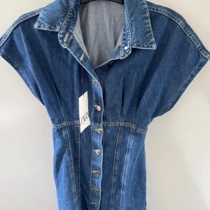 Jeansklänning med knappar från Zara - Snygg jeansklänning från Zara i klassisk blå denim. Klänningen har korta ärmar, markerad midja och knappar hela vägen framtill. Perfekt krage och cool, avslappnad passform. Enkelt att styla med sneakers eller boots för en trendig look.