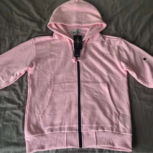Ljusrosa hoodie från Stone Island - Säljer en ljusrosa hoodie från Stone Island med dragkedja framtill och klassisk huva. Tröjan har ribbade muddar, två stora fickor och en diskret logga på ärmen. Perfekt för dig som gillar streetwear och vill sticka ut med en snygg färg.