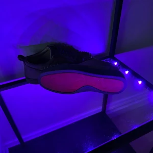 Louboutin  - Säljer ett par svarta sneakers från Christian Louboutin i mocka med ikoniska röda sulor. Skorna har snörning och är dekorerade med svarta nitar över tån för en edgy look. Perfekta för dig som vill sticka ut med exklusiva detaljer och lyxig känsla.