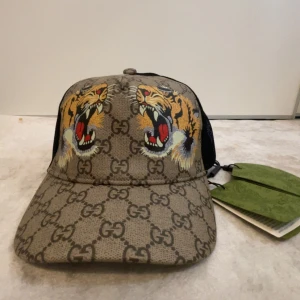 Gucci keps med tigertryck och GG-mönster - Snygg keps från Gucci med klassiskt GG-monogram i beige och brunt, prydd med två färgstarka tigrar framtill. Baksidan är i svart mesh för extra ventilation och justerbar passform med grön-röd randig detalj. Perfekt statement-piece för dig som vill sticka ut.