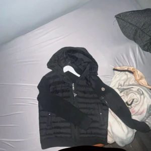 Svart pufferjacka med huva från Moncler - Säljer en svart pufferjacka från Moncler med stickade ärmar och huva. Jackan har dragkedja framtill och två fickor med knappar. Perfekt för kyliga dagar och riktigt snygg till streetwear-stilen. Kan ta emot byten men endast i storlek s-xs