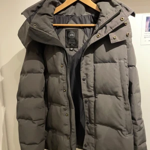 Canada goose Whyndham - Säljer en grå pufferjacka från Canada Goose i storlek M. Jackan har en rejäl huva, dragkedja och knappar framtill samt stora fickor. Klassisk quiltad design och patch med logga på ärmen. Perfekt för kalla dagar när du vill hålla stilen.