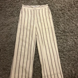 Randiga linnebyxor från Joelle - Snygga beige linnebyxor från Joelle med raka, vida ben och vertikala bruna ränder. Byxorna har hög midja, knappstängning och sidfickor. Perfekta för en chill och trendig look under varma dagar.