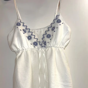Supersöt vit topp med blå broderi - Superfin vit topp med tunna axelband och blått blommigt broderi vid urringningen. Den har knytband i midjan. Säljer på grund av den endast har använts ett fåtal gånger 💞🙌🤩
