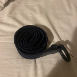 Mörkblå flätat bälte med läderdetaljer - Säljer ett mörkblått flätat bälte med snygga läderdetaljer vid spännet. Bältet har en klassisk design och passar perfekt till jeans eller chinos. Spännet är i metall och bältet är flexibelt tack vare det flätade materialet.