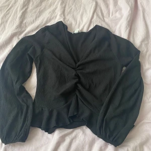 Svart omlottopp med volangdetalj - Säljer en svart omlottopp med långärmade puffärmar och snygg v-ringning. Toppen har en rynkad mitt och volangkant nedtill som ger en cool och feminin vibe. Materialet är lätt och har en krinklad struktur som känns skönt mot huden.