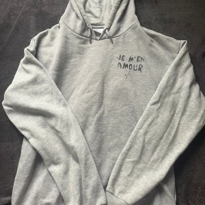 Grå hoodie från Gina Tricot med tryck🤍 - Snygg grå hoodie från Gina Tricot med svart tryck 'JE M'EN AMOUR' och en stjärna på bröstet och ryggen. Tröjan har huva med snörning och är tillverkad i mjuk bomullsmix. Perfekt för chill dagar och streetstyle. Den har en liten fläck, se bild 5 och den är lite nopprig men det är inte något man tänker på enligt mig🫶🏻