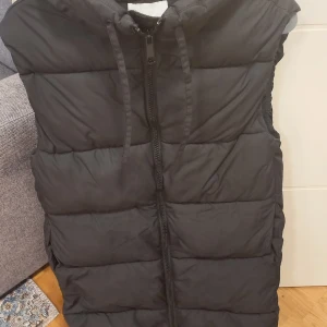 Svart dunväst från H&M XS - Svart dunväst från H&M i storlek XS lite overzise.Västen har huva med dragsko. Materialet är polyester och västen är lätt vadderad för extra värme.